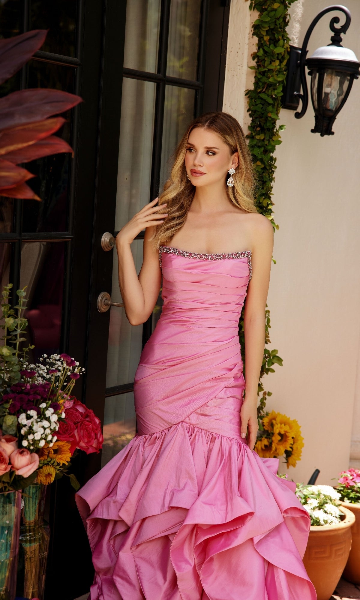 A woman in the Ava Presley 40068 Strapless Ruffled Mermaid Prom Dress stands by a glass door, surrounded by colorful flowers and greenery.